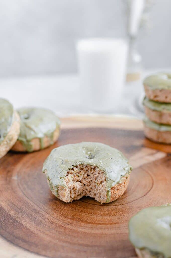A matcha glazed donut with a bite taken out of it and other donuts scattered around with a glass of milk in the background.