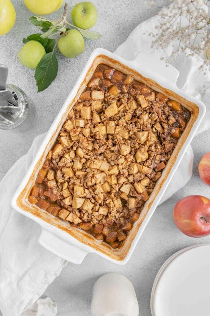 Vegan baked apple oatmeal in a baking dish surrounded by apples.