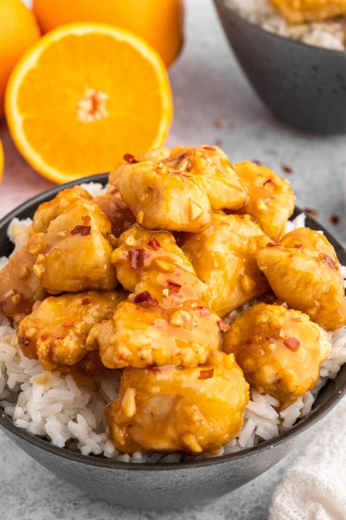 a black bowl of orange chicken and white rice next to fresh oranges