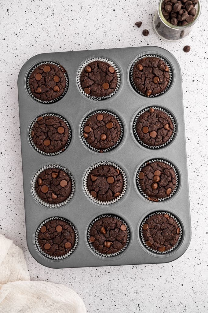 Protein chocolate muffins in a muffin tin next to a glass jar of chocolate chips.