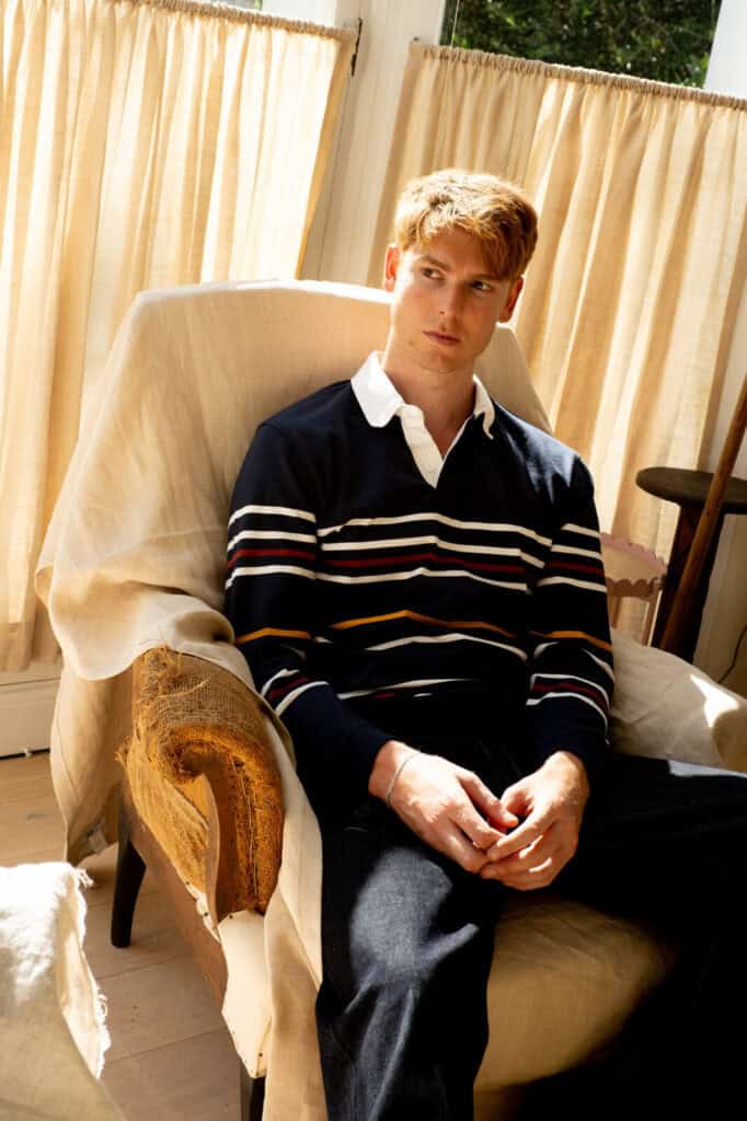 Dan Whitlam, a young man with red hair, sits on a worn, fabric-covered armchair in a sunlit room with beige curtains, wearing a navy sweater with white, yellow, and red stripes over a white collared shirt and dark pants.