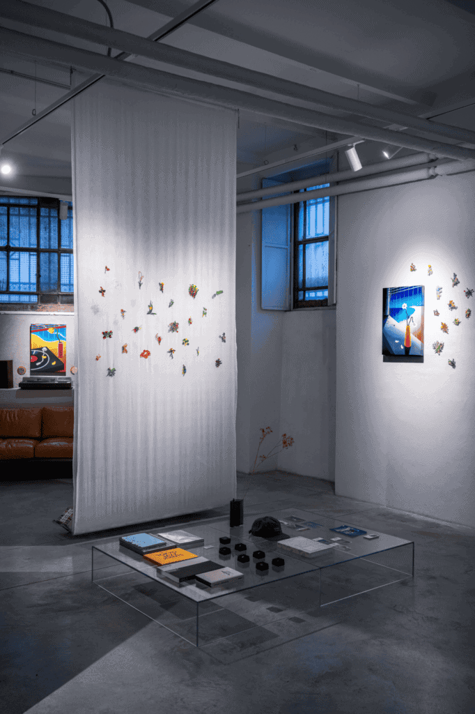 A modern art gallery interior inspired by Hiroki Yagi, featuring sheer white fabric hanging from the ceiling with colorful origami pieces. Artworks adorn white walls, while a glass coffee table with books sits on the concrete floor.