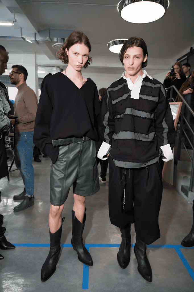 Two models stand indoors. The model on the left wears a black top, green leather shorts, and black boots. The model on the right wears a black and gray textured sweater, loose black pants, and black boots. People converse in the background.