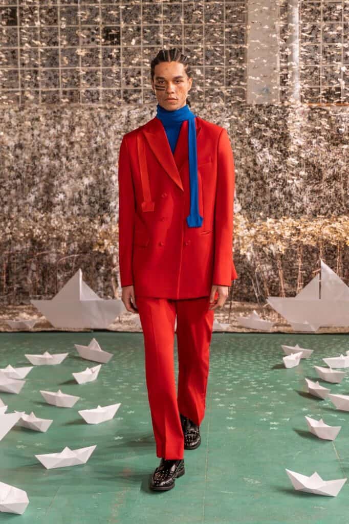 A person in a bright red suit with a blue turtleneck and scarf walks on a green floor scattered with white paper boats, in front of a backdrop with metallic, reflective decorations and geometric glass panes.