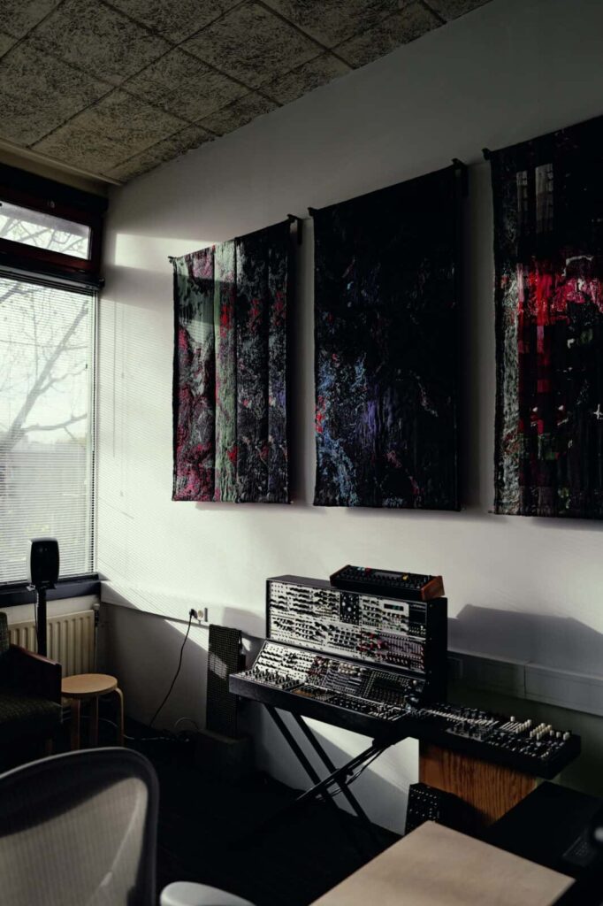 A music studio corner with a large modular synthesizer on a stand beneath three abstract wall hangings. Sunlight streams through a window, illuminating part of the room. A stool and chair are also visible, Artworks by Ben Kreukniet.