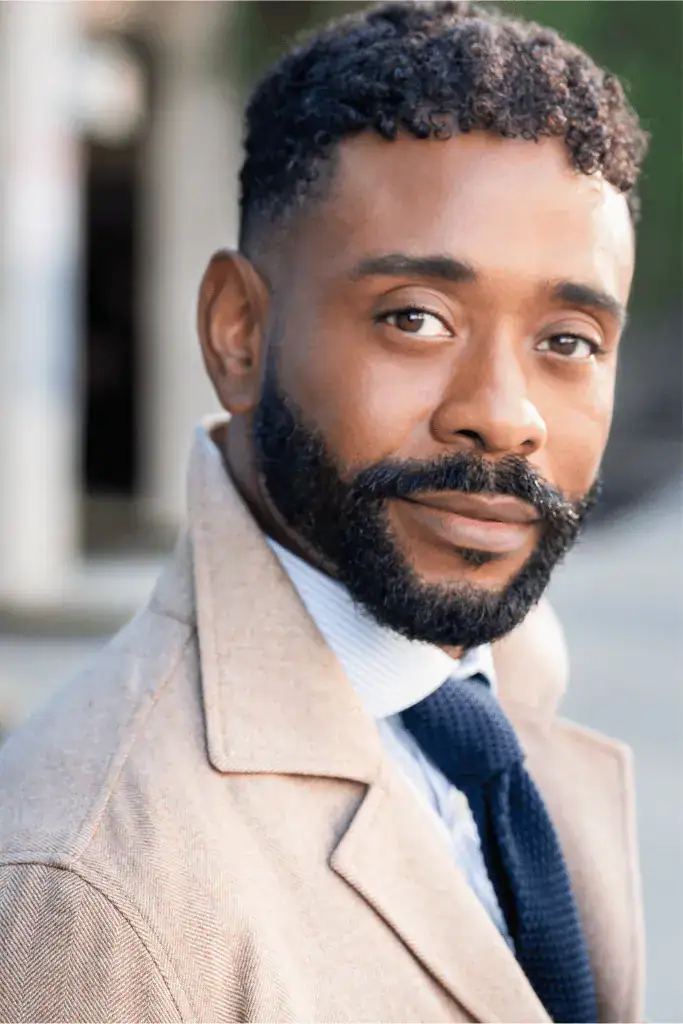 Photo of James Brown III, a brown-skinned man with black, short hair and a beard and moustache wearing a tan sport coat with dark tie, gazing at the photographer with a neutral expression. 