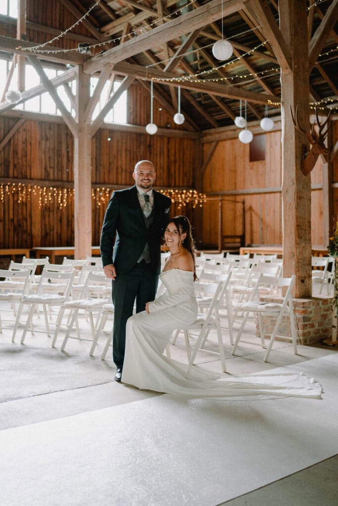 Eine Braut in einem weißen Kleid sitzt lächelnd neben einem stehenden Bräutigam im dunklen Anzug im Projektraum Aumühle. Der rustikale, aus Holz gefertigte Veranstaltungsort ist wunderschön mit Reihen weißer Stühle und Lichterketten geschmückt, die von der Decke hängen und eine bezaubernde Atmosphäre schaffen.