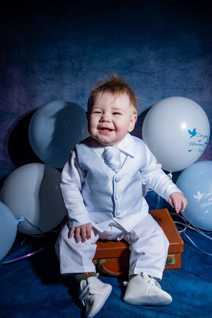 Smiling child in formal white outfit seated on suitcase, surrounded by balloons — part of EJC Photography’s joyful, bespoke portrait sessions for special occasions.