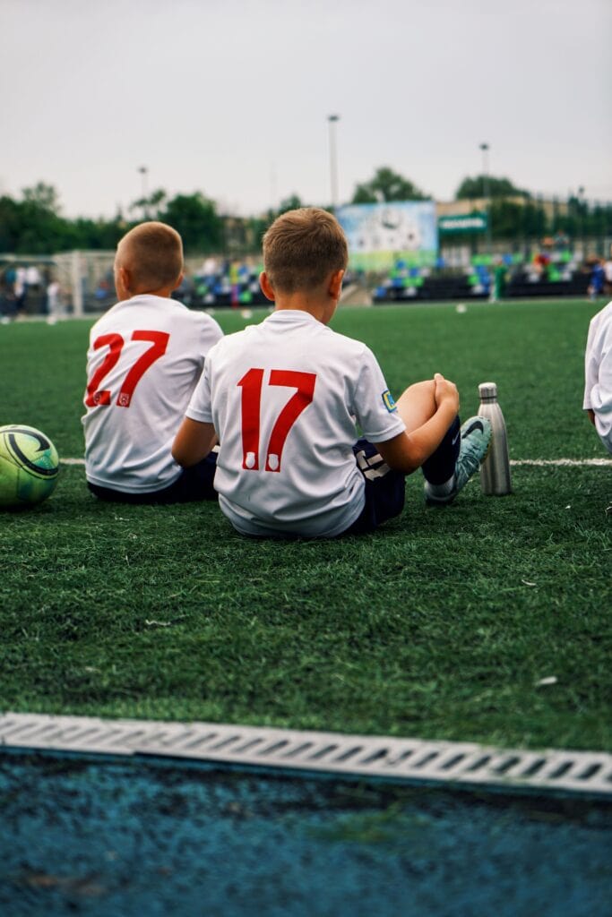 Boys in soccer jerseys sitting on a turf field during a summer game.