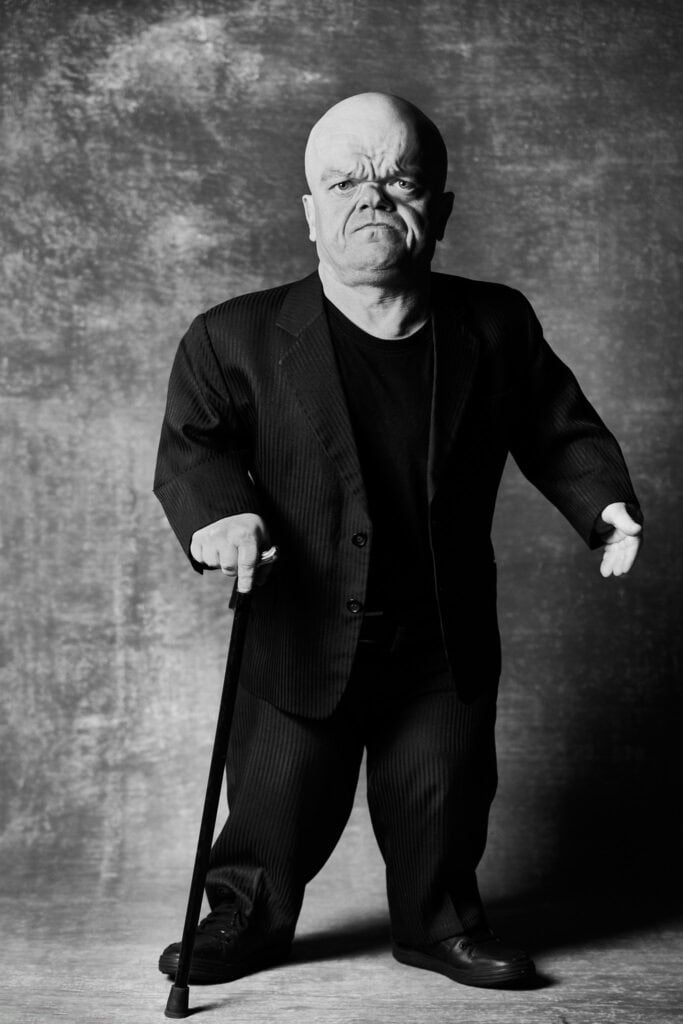 Portrait de l'acteur Belge Chris Willemsen debout, en costume, appuyé sur une canne