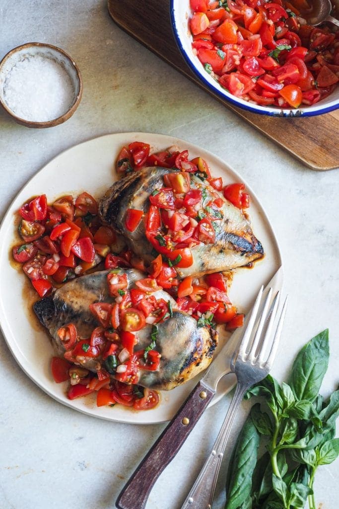 Paula's Apron Pechugas de Pollo Caprese Esta receta de Pechugas de Pollo Caprese es una deliciosa variación de la clásica ensalada italiana! Primero se marina el pollo con un adobo balsámico, para que quede más tierno. Luego se asa a la parrilla, donde el vinagre balsámico se carameliza. Finalmente, se cubre con queso mozzarella y una deliciosa ensalada de tomates cherry con albahaca. Aquí se puede ver un plato con dos pechugas de pollo cocinadas, la mozzarella y la ensalada de tomate por encima. A bajo a la derecha hay un tenedor y un cuchillo apoyados al plato y un manojo de albahaca. Arriba a la izquierda hay un pequeño bol con sal y arriba a la derecha un bol con extra de ensalada encima de una tabla de madera.