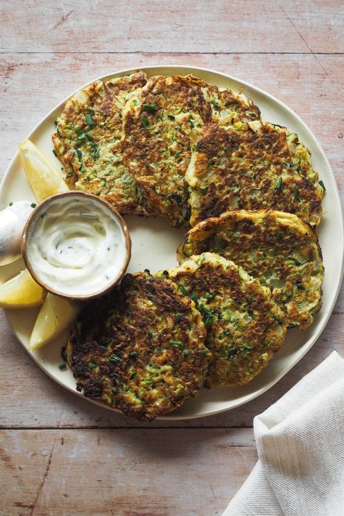 Paula's Apron Easy Courgette Fritters (Gluten Free) These Easy Courgette Fritters with Lemony Herby Crème Fraîche are a game changer for weeknight meals. They are gluten-free and combine eggs, courgette, and a delicious mix of herbs and spices that make them yummy while being light. In this overhead shot, the fritters can be seen placed on a plate in a semi circular fashion around a little bowl with the crème fraîche dip. A piece of a kitchen cloth can be seen on the bottom right corner.