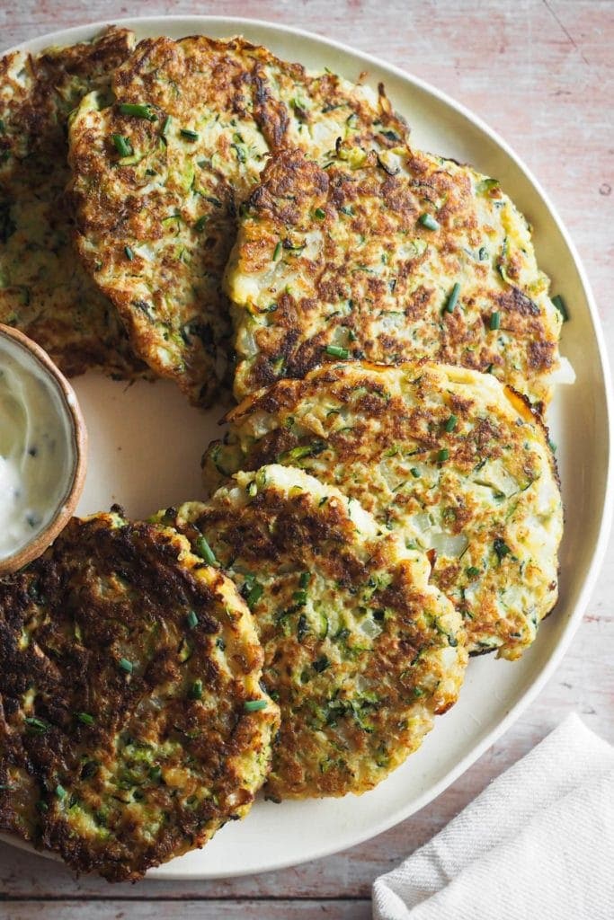 Paula's Apron Easy Courgette Fritters (Gluten Free) These Easy Courgette Fritters with Lemony Herby Crème Fraîche are a game changer for weeknight meals. They are gluten-free and combine eggs, courgette, and a delicious mix of herbs and spices that make them yummy while being light. In this close up overhead shot, the fritters with all the details of the texture can be seen placed on a plate in a semi circular fashion around a little bowl with the crème fraîche dip. A piece of a kitchen cloth can be seen on the bottom right corner.