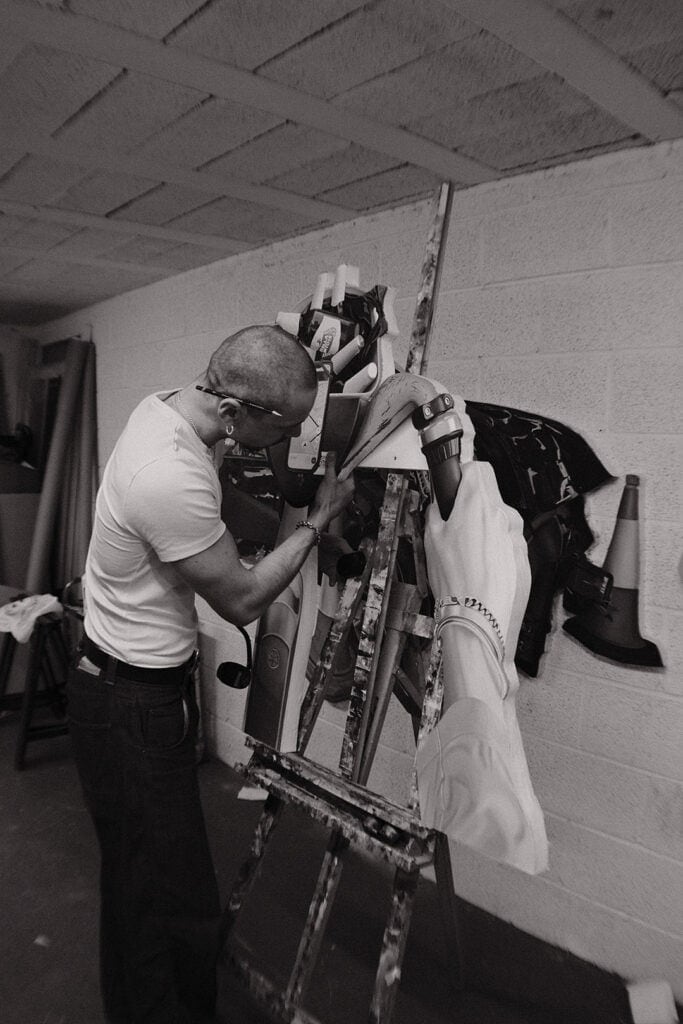 Callum Eaton with a shaved head and glasses, works on a painting at an easel in a studio, depicted in black and white. The artwork shows a large hand holding an object as the person intently concentrates.