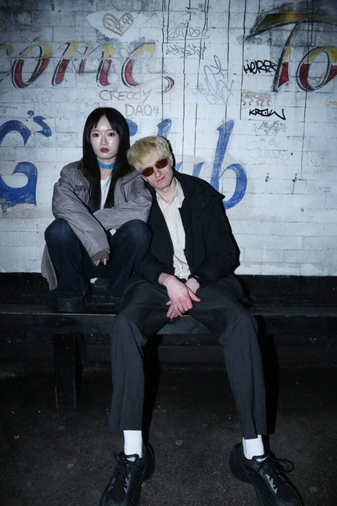 Two people sit on a bench in front of a white brick wall covered in graffiti. One person wears a gray jacket and has long dark hair, while the other wears sunglasses, a light shirt, and dark pants. Both look directly at the camera.