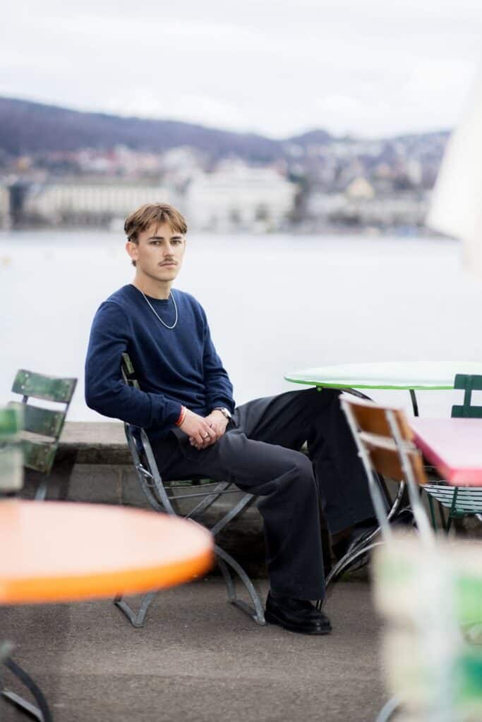 A portrait of author Nelio Biedermann sitting at an outdoor café table with a blurred city and lake background, wearing a dark blue sweater and grey trousers.