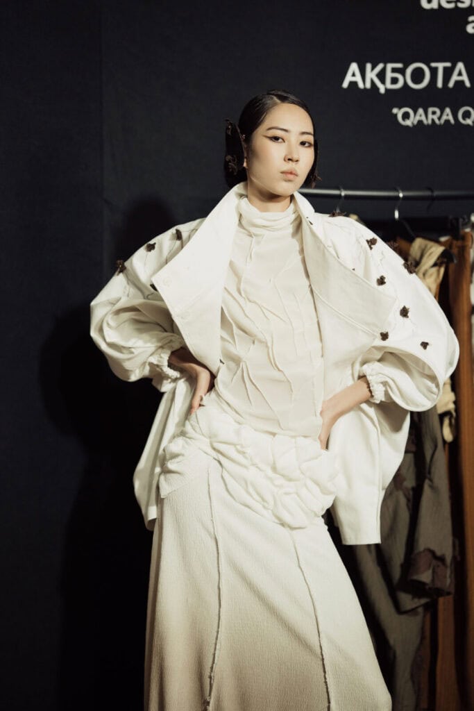 Model wearing Akbota Kapsalan’s white "Qara Qandyagash" collection, featuring a textured high-neck top and a jacket adorned with small dark floral accents at Visa Fashion Week Tashkent.