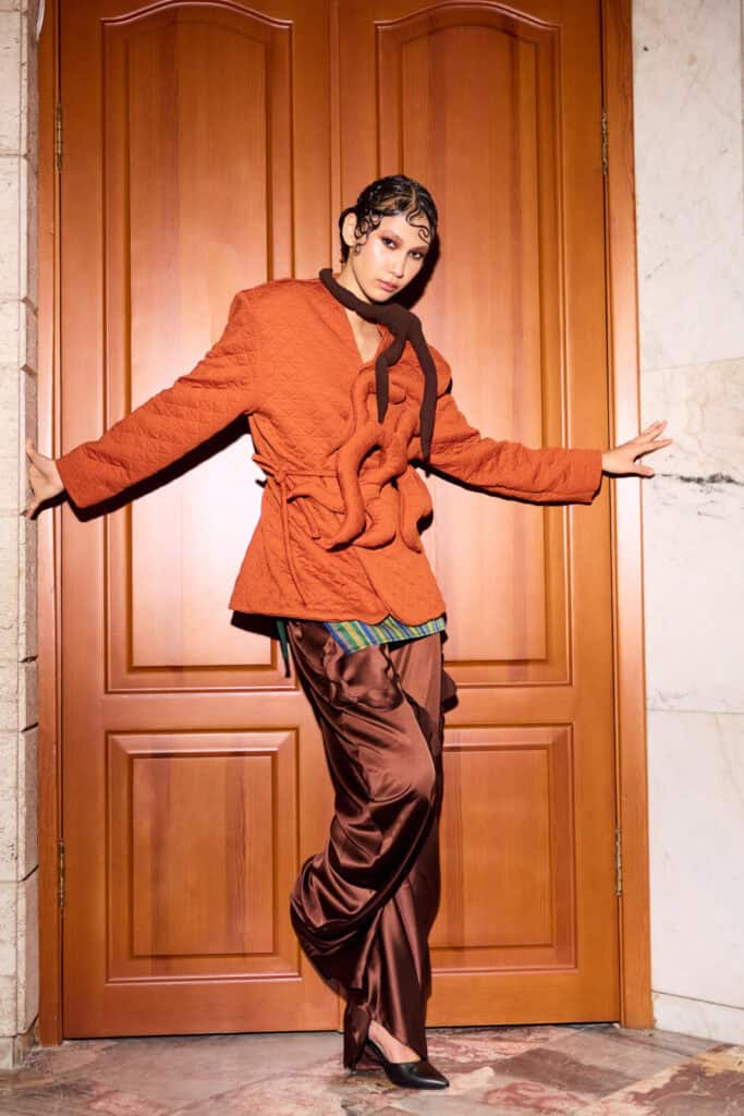 A model showcasing a No.sugar ensemble featuring an orange quilted blazer with 3D root details and flowing brown satin trousers against a classic wooden door backdrop.