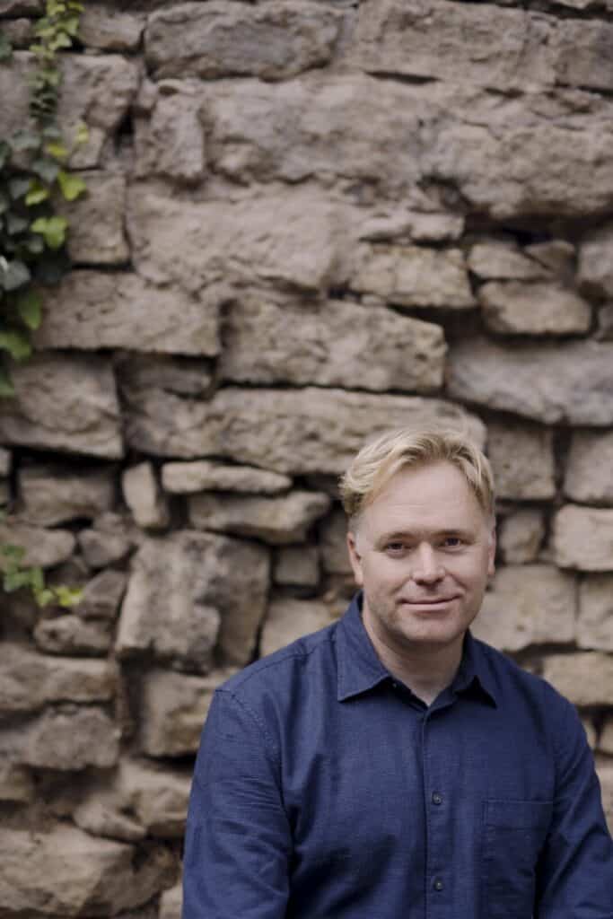 Graeme Richardson, author of the poetry collection Dirt Rich, posing in a blue button-down shirt in front of a rustic stone wall with green ivy.