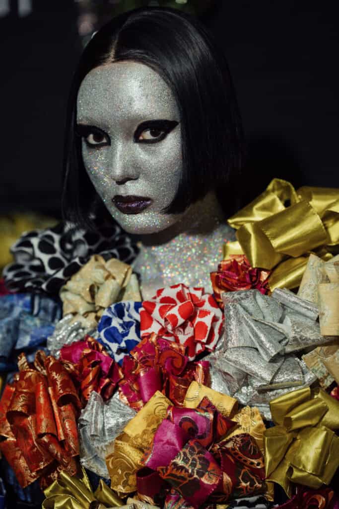 Close-up of a model with silver glitter makeup and dark purple lipstick wearing a top made of ornate gift bows, part of the "Fake Prada" narrative by Esbolat Esmukhanov.