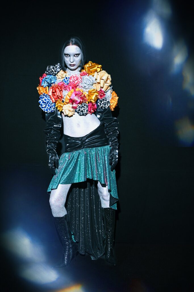A model in Esbolat Esmukhanov’s collection featuring a bodice made of multicolored gift bows, metallic green skirt, and silver glitter body paint at Almaty Fashion Week.