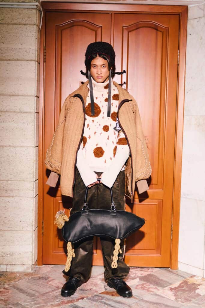 A full-length shot of a "Kökpar" collection look, including a quilted tan jacket, faux fur spotted sweater, and a black leather bag with rope legs at Visa Fashion Week Almaty.