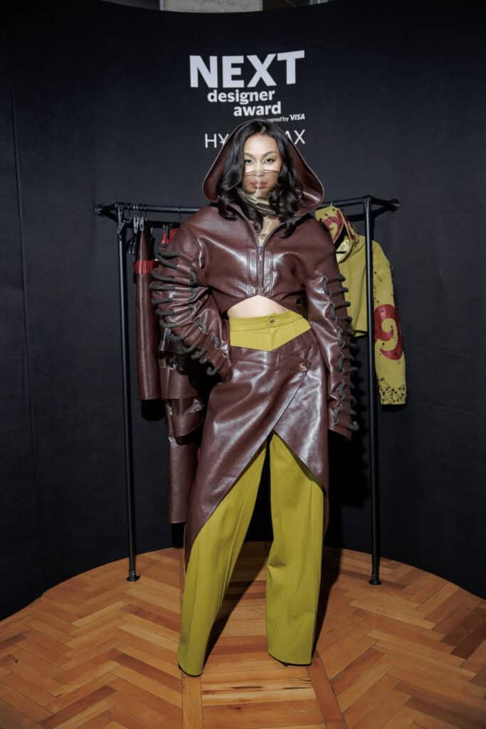 A full-length view of SAKH's "Tamyr" look, featuring a maroon leather wrap skirt and mustard trousers on the NEXT Designer Award stage in Almaty.