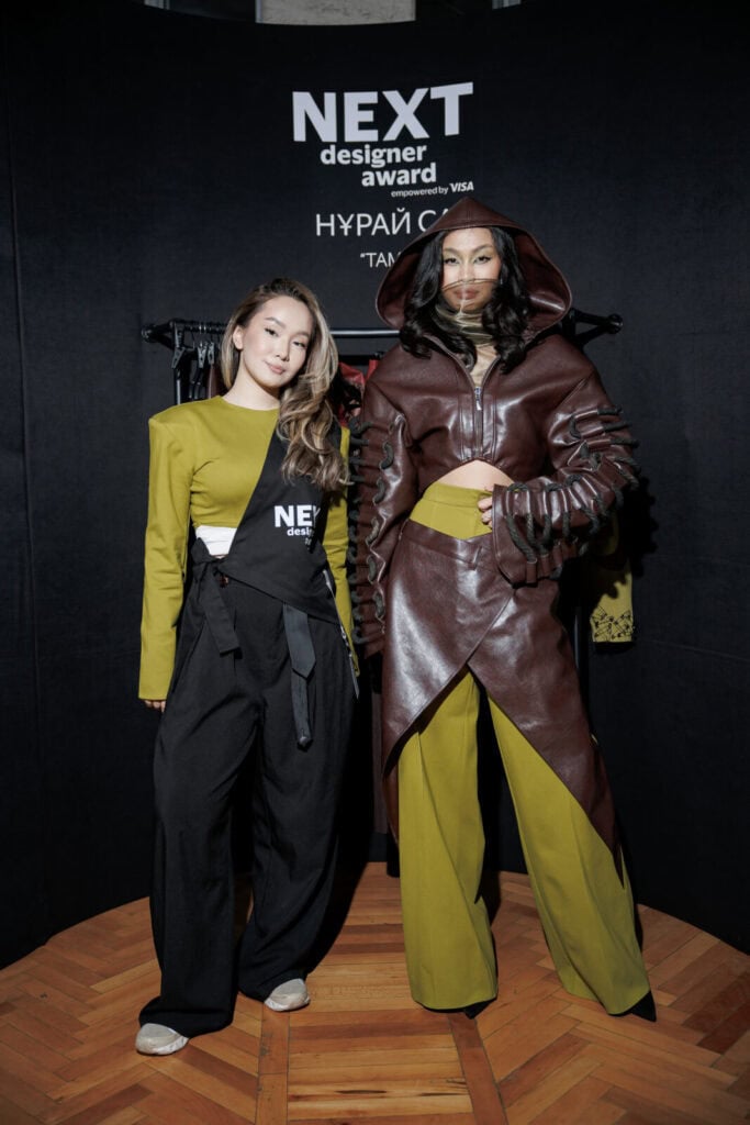 Designer Nuray Sakh posing with a model in her "Tamyr" collection at Visa Fashion Week Almaty, showcasing a fusion of nomadic roots and modern leatherwork.