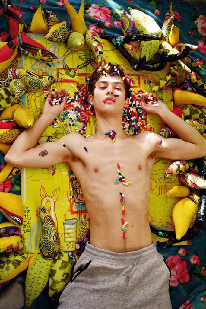 A shirtless young man lying on his back amid colourful printed fabric and stuffed toys, rainbow sprinkles scattered across his chest and in his hands, eyes closed — photograph by Slava Mogutin