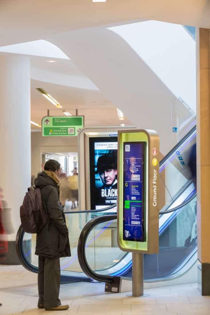 Escalator index sign located at the base of an escalator. The lozenge shaped sign has a green acrylic infill and details about the various amenities on each floor are listed in a blue panel. In the distance is a directional prompt sign, suspended from the ceiling similarly with a green acrylic infill and white text on a darker green background