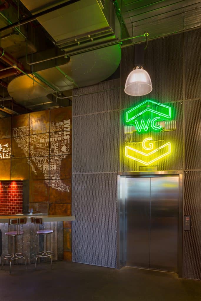 Stepped back view of the neon index sign. Installed on wire mesh panels above the lift door, the sign features a green outline of an arrow head pointing upwards with WC written below. Stacked underneath is a letter G in yellow neon and a downward pointing arrow head