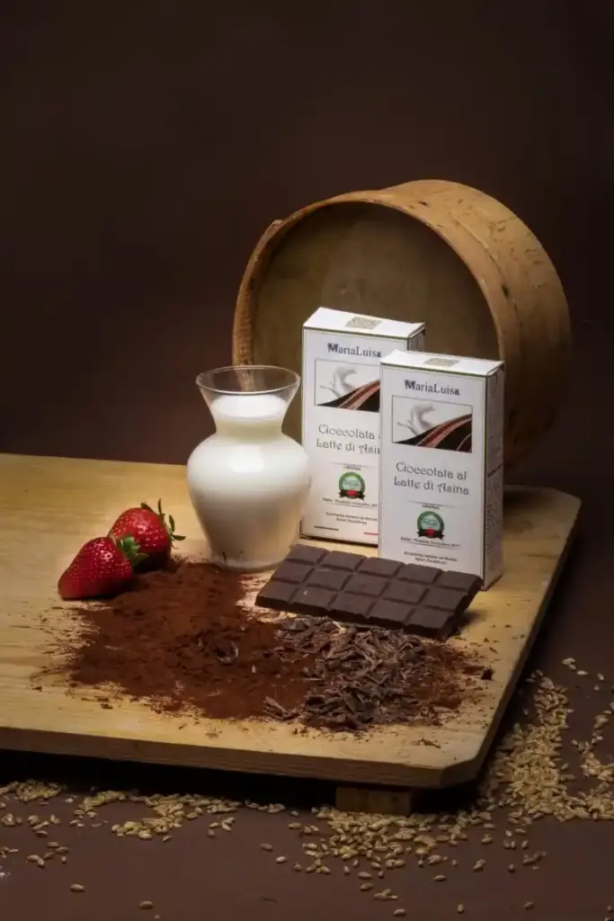 Cioccolato fondente con latte di asina Marialuisa su una tavola di legno con confezione, giara di latte di asina fresco, polvere di cacao, fragole e setaccio in legno