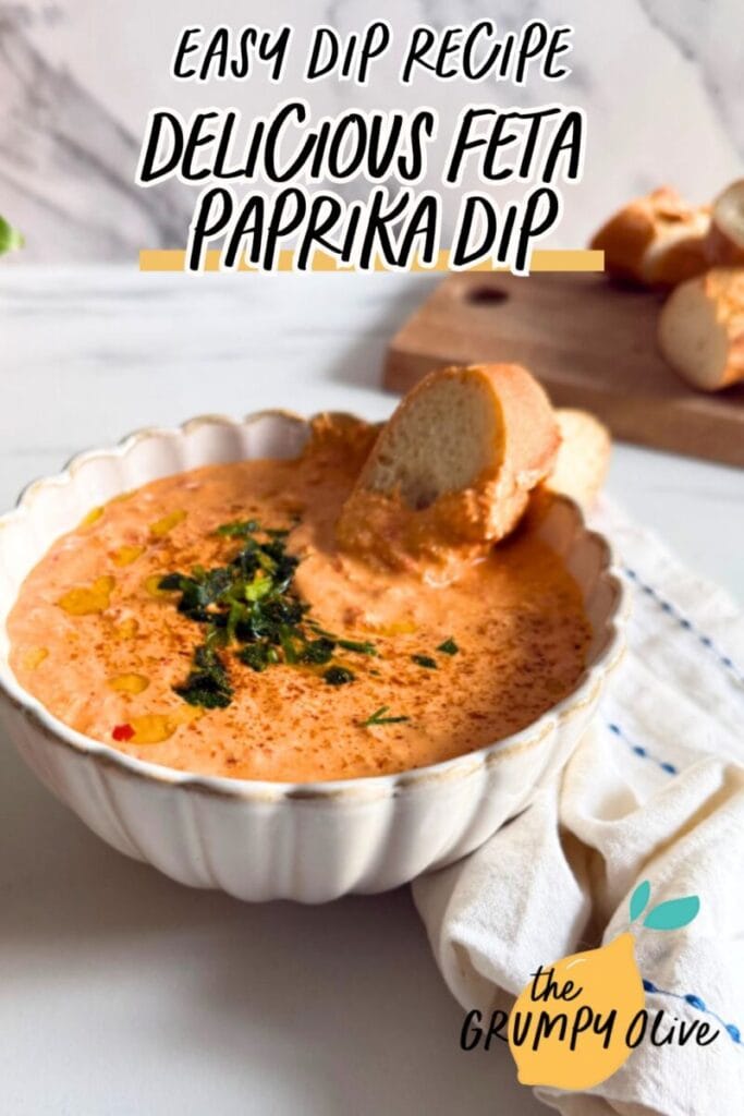 Feta Paprika Dip - Easy & Quick Dip Recipe 11 pin image paprika dip in a bowl with a slice of bread, more bread on a wooden board behind and some on the side, a towel, a plant, on a marble background