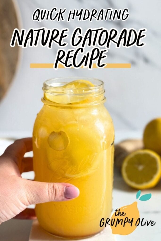 Easy 5 Minutes Homemade Natures Gatorade Recipe 9 pin image hand holding a jug of natures gatorade on a marble coaster, straws on front, lemons and ginger in the back with a bowl of salt