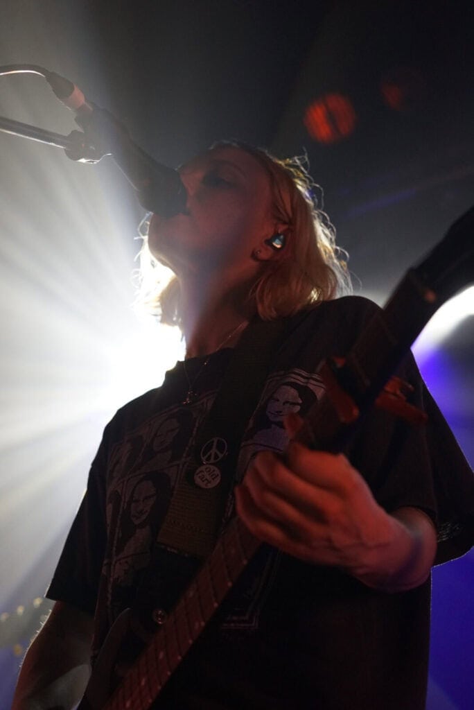 A musician with shoulder-length blonde hair from the Momma band sings into a microphone and plays electric guitar on stage, illuminated by dramatic backlighting that creates a striking silhouette effect.