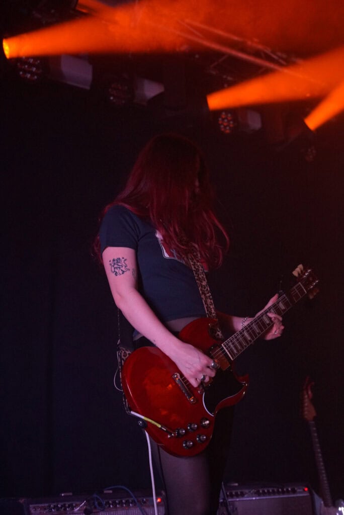 A musician with long red hair from the Momma band plays a red electric guitar on stage under orange lights, wearing a dark t-shirt and displaying a tattoo on their upper left arm. Amplifiers and equipment are visible against the dark background.