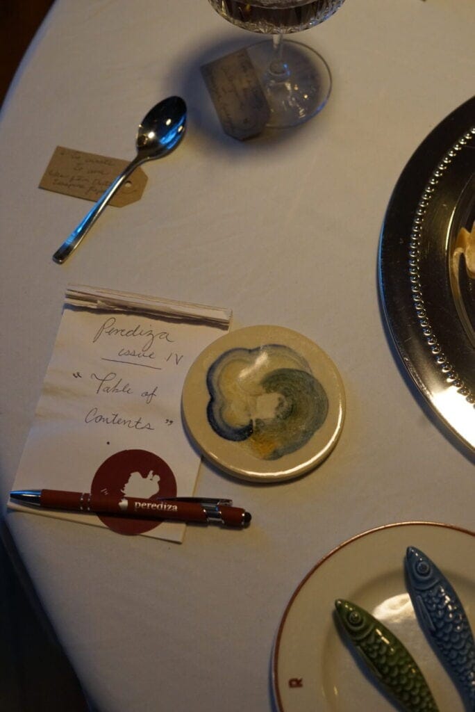 A notepad labeled Porediza Castle IV Table of Contents sits on a table with a red pen, a round ceramic coaster, a spoon, a glass of wine, and fish-shaped chopstick rests on a white tablecloth.
