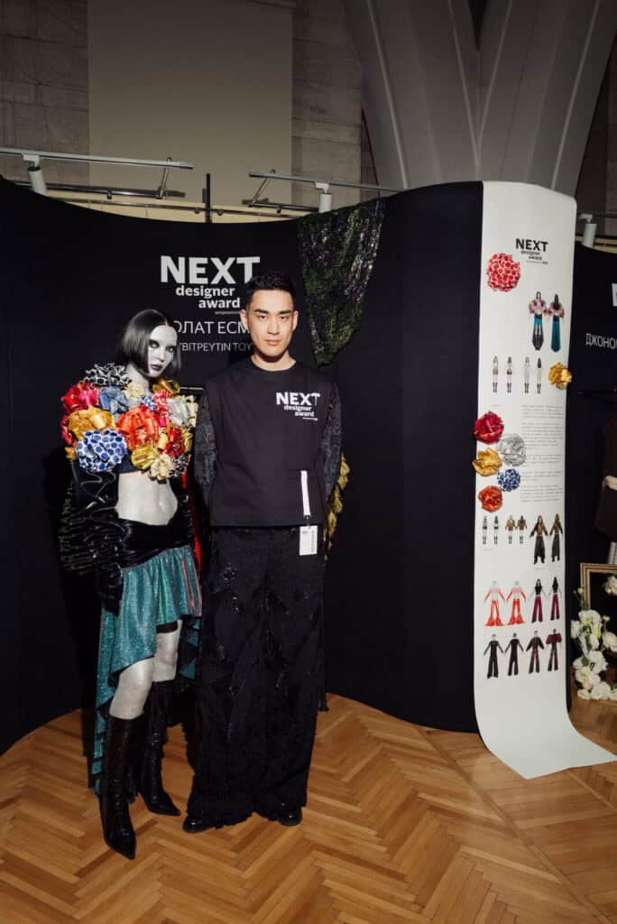 Winner Esbolat Esmukhanov standing next to his model in a booth at Visa Fashion Week Almaty, showcasing his award-winning designs that critique mass-market fashion culture.