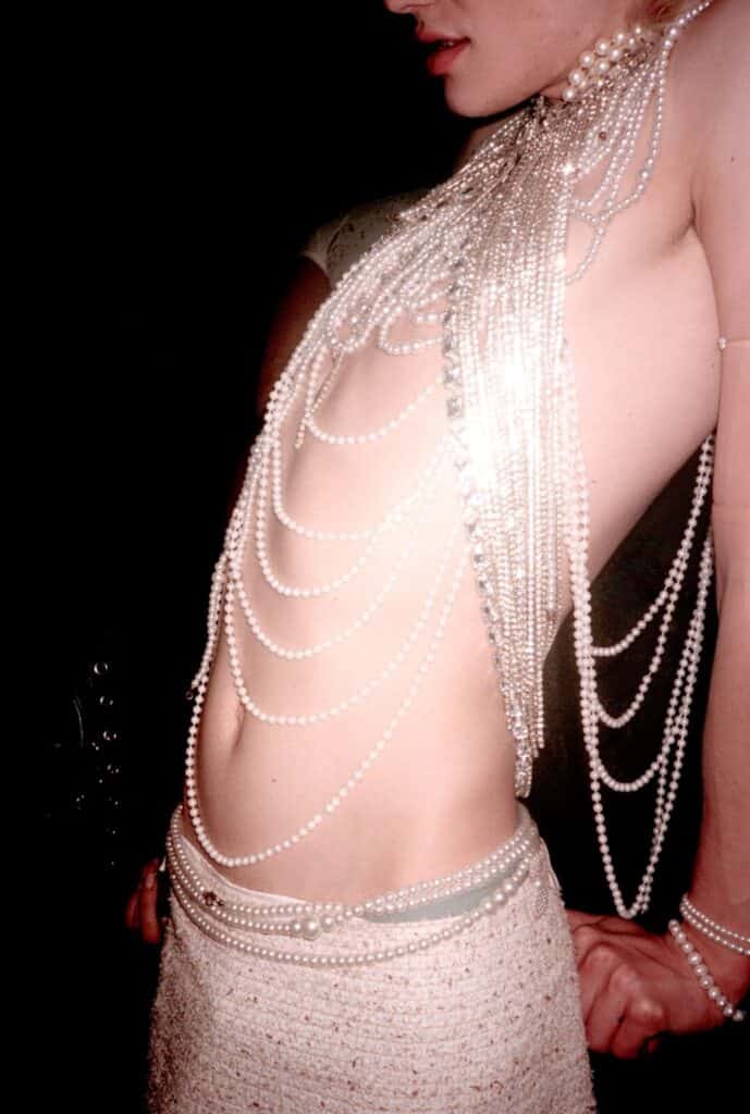 Side profile of a figure draped in cascading pearl and crystal necklaces against a black background, torso bare beneath layered strands of white beads falling to a textured skirt — photograph by Arthur Samier