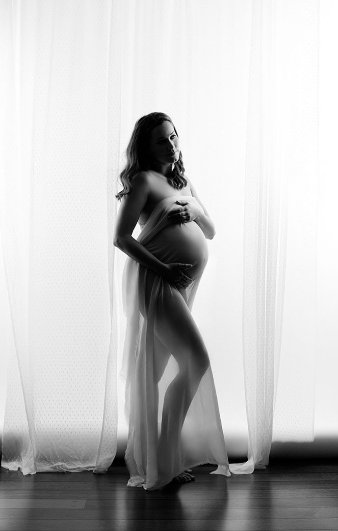 Elegant gravidfoto av en gravid kvinne foran et lyst gardin, fokus på skjønnhet og graviditet, ideell for gravidfoto, morskap og spesielle anledninger.