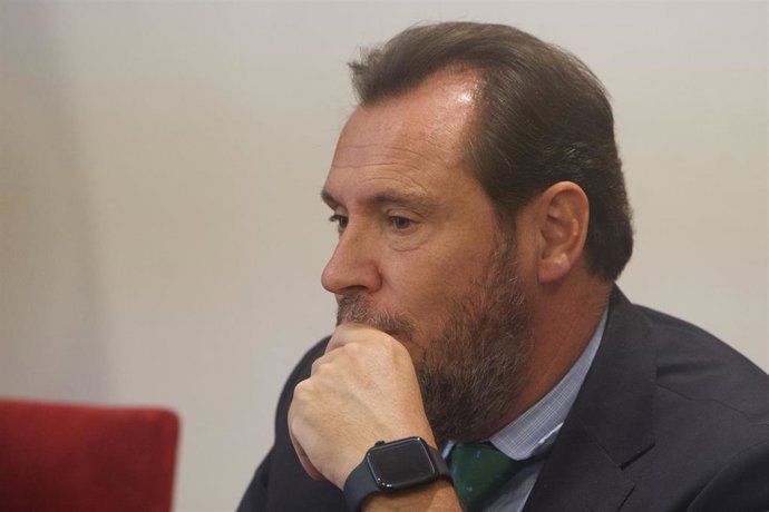 Hombre con barba y cabello castaño, pensativo, sentado en una mesa con fondo blanco y una silla roja, en Madrid.