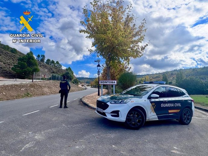 Un agente de la Guardia Civil con uniforme, un coche patrulla y un letrero que indica la entrada al pueblo de Hontoba.