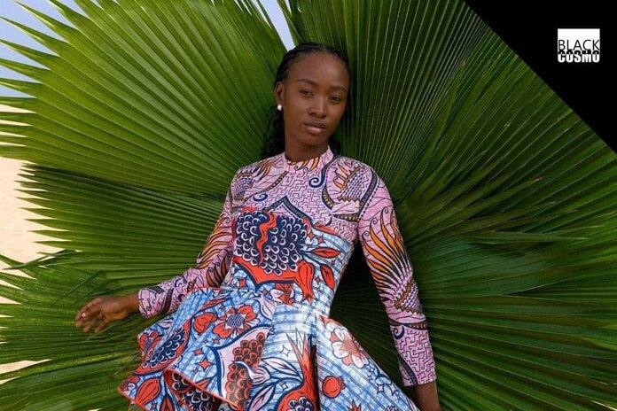 A model in a wax print dress stands behind a large, green plant.