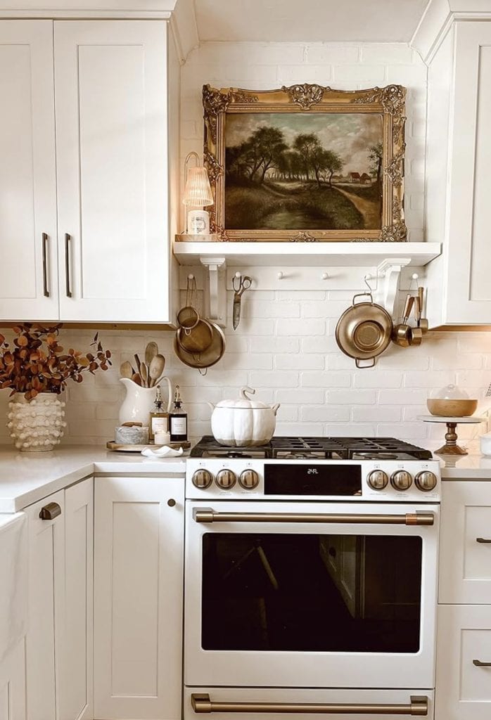 White kitchen and stove decorated with brass pots, pans, and a pumpkin kettle — cozy fall‑inspired top of stove decor ideas from By Paige Nicole.