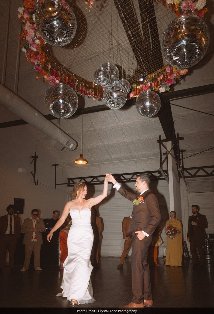 This image highlights a bride and groom enjoying their first dance, while surrounded by their wedding party. The image is set in a white modern industrial warehouse, and the dancing is under a decorative disco ball installation, hanging from the rafters. The bride and groom are smiling at one another and holding hands, the bride in a long white dress and the groom in a brown suit.