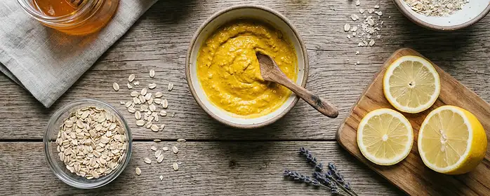 A flat lay of natural skincare ingredients like honey, turmeric, lemon, oats, and a bowl with a yellow face mask mixture on a wooden surface