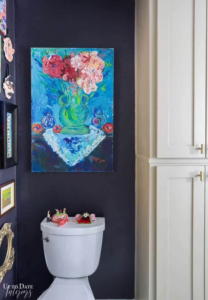 Bold blue floral large painting over toilet and next to tall taupe cabinets. Black wall paint.