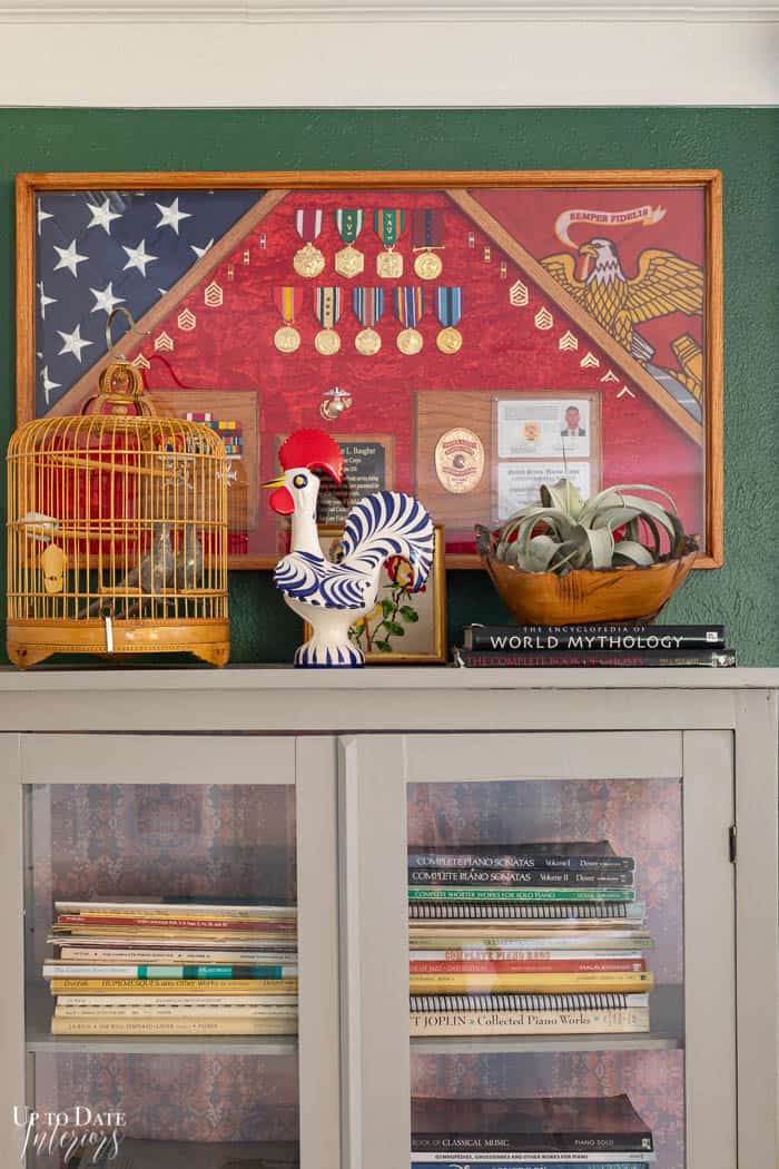 Military wall art with flag and ranks, ribbons on top of cabinet with bamboo bird cage, rooster, books, and air plant.