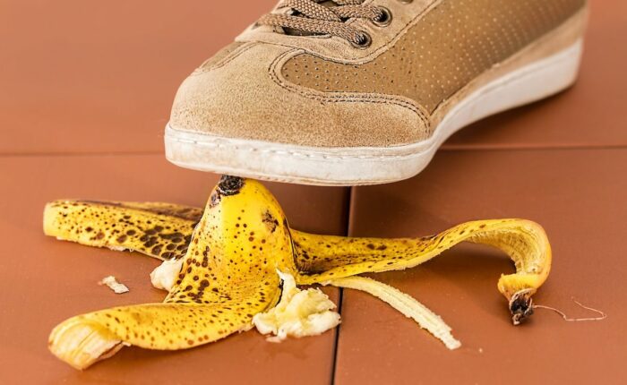Schuh tritt auf eine braun gesprenkelte Bananenschale auf einem braunen Fliesenboden, symbolisiert eine Gefahrensituation.