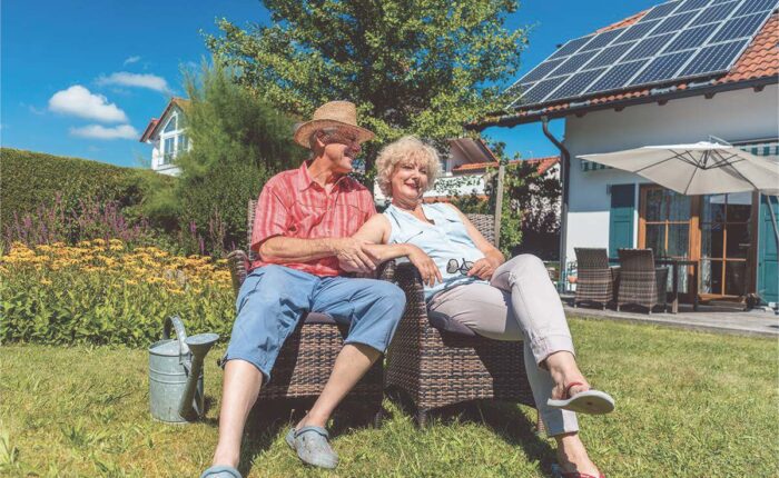 Seniorenpaar sitzt entspannt im Garten vor einem Haus mit Solaranlage bei sonnigem Wetter.